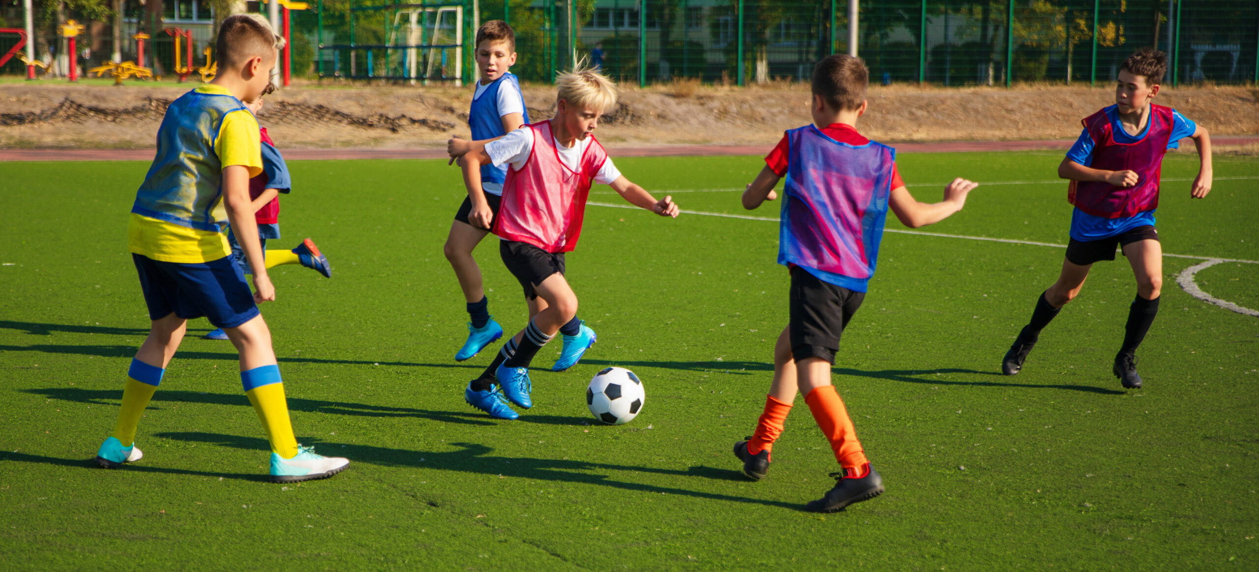 The photo shows a group of young soccer players in colorful jerseys on a green turf field. They are actively playing and competing for the ball, with some players in motion and others watching.